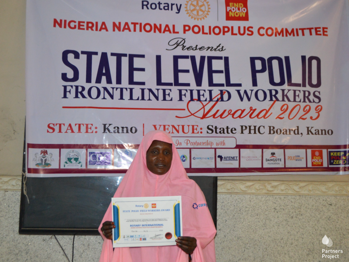 Zainab in a pink khimar holds her award in front of a Rotary banner that says State Level Polio frontline field workers award 2023